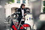A man with long hair and a beard at a bike rental station starts the hiring process with his cell phone. In the background, house facades.