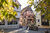 A small square in the old town of Bad Cannstatt in Stuttgart can be seen. In the center is an old fountain, in the foreground are autumnal deciduous trees and in the background is an old half-timbered house.